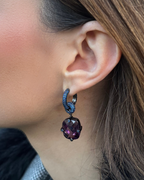 Pendientes Lyss en plata de primera ley 925 con baño de rodio negro. Diseño sofisticado compuesto por un aro engastado con piedras de zafiro azul y una piedra central en corte Radiant Ceilán de color violeta, sujeta por cuatro garras decoradas con pequeños zafiros. Joya elegante y exclusiva con un toque de misterio.